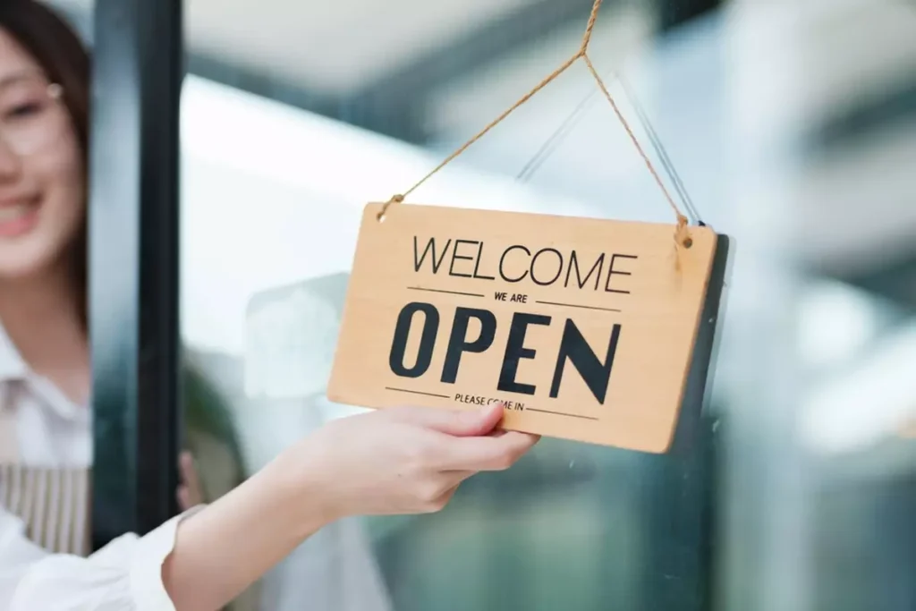 A shopkeeper flipping over a sign hanging in her front door that reads 'WELCOME we are OPEN.'