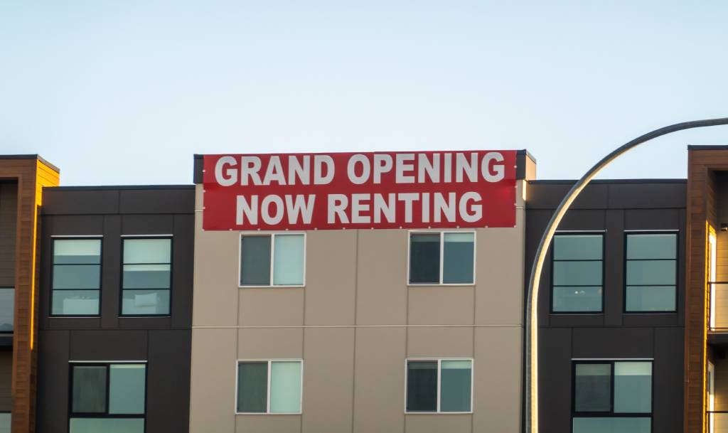 Exterior view of a modern apartment building featuring a large red banner with white text that reads Grand Opening Now Renting.
